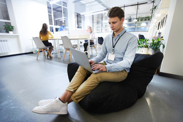 Serious concentrated handsome hipster manager with conference badge in neck sitting in bean bag and...