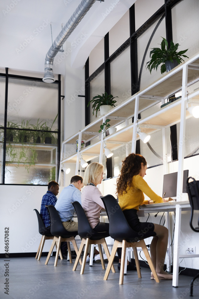 Wall mural Group of busy concentrated creative colleagues in casual clothing sitting at tables with devices and working with papers in modern office