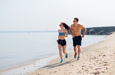 fitness, sport and lifestyle concept - happy couple in sports clothes and sunglasses running along summer beach