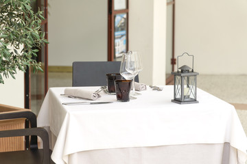An empty table for two in a restaurant, covered with a white tablecloth with two table sets and a lantern, in a cafe or restaurant outside.