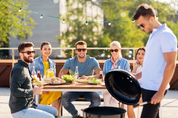leisure and people concept - happy man grilling on bbq and friends at rooftop party