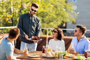 leisure and people concept - happy party host offering meat to his friends at bbq on rooftop in summer