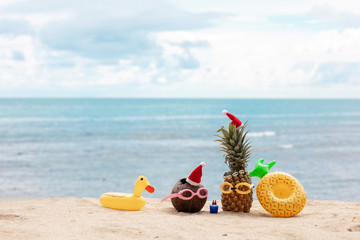  funny attractive pineapples and coconut in stylish sunglasses on the sand against turquoise sea. Wearing christmas hats. Christmas and new year vacation concept on tropical beach.