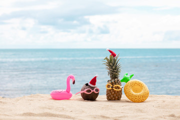  funny attractive pineapples and coconut in stylish sunglasses on the sand against turquoise sea. Wearing christmas hats. Christmas and new year vacation concept on tropical beach.