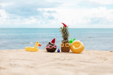  funny attractive pineapples and coconut in stylish sunglasses on the sand against turquoise sea. Wearing christmas hats. Christmas and new year vacation concept on tropical beach.