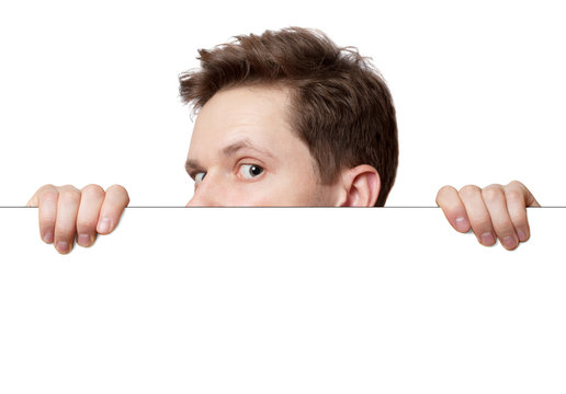 Young man with surprised eyes peeking out from behind billboard paper poster. Man peeking out from the edge and looking at camera isolated on a white background