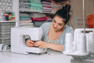 Portrait of young woman seamstress sitting and sews on sewing machine. Tailor making a garment in her workplace. Hobby as a small business concept