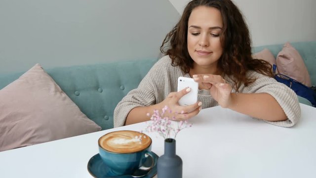 Attractive woman take mobile photo of cappuccino at cafe. Curly brunette use smartphone at modern cafe.