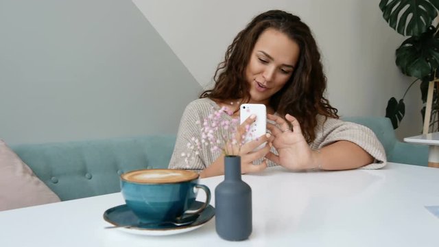 Attractive woman take mobile photo of cappuccino at cafe. Curly brunette use smartphone at modern cafe.