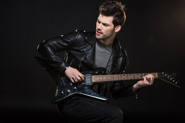 handsome rocker in leather jacket playing electric guitar and looking away isolated on black