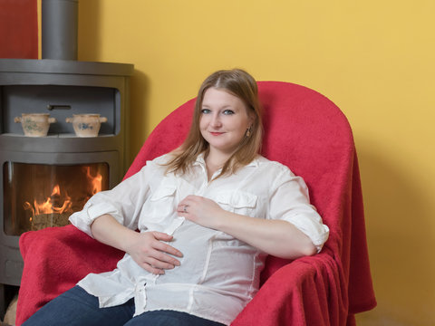 Smiling Pregnant Young Woman Is Relaxing Sitting In Chair At Burning Fireplace Stove In Home. The Woman Is Looking At The Camera.