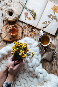 From above hands of unrecognizable lady holding bunch of wild flowers while lying on thick soft blanket near tea with honey and notebook with dried plants