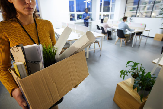 Serious sad woman in sweater carrying cardboard box full of stuff and leaving office after dismissal