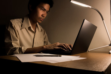 A busy business man is sitting on the chair for working overtime at office at night. He use a led lamp for lighting his desk for writing and typing laptop for research information