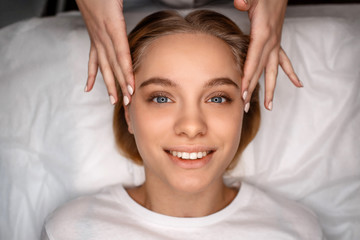 Cheerful attractive young woman lying on white couch. Beautician touch her face with bare hands. Model smile and look straight.