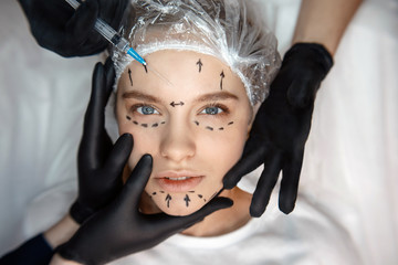 Calm peaceful young woman lying on couch and look straight forward. Her face is marked. Three hands touching face skin. Fourth hold syringe for injection. - obrazy, fototapety, plakaty