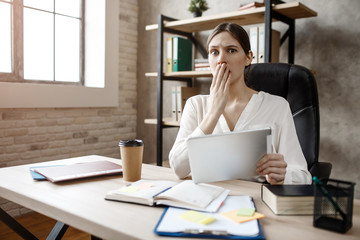 Young scared buisnesswoman in room at table. She hold tablet and cover mouth with hand. Model look straight forward.