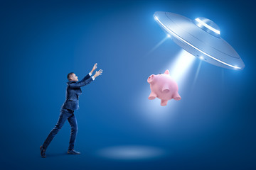 A businessman raising hands toward a UFO which is pulling a piggy bank toward its open hatch by some invisible force.