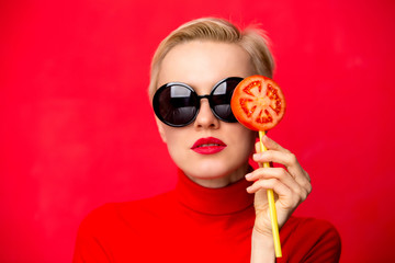 Summer fashion woman wearing sunlglasses and holding tomato lollipop against red wall. Beauty,...