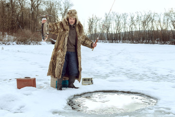 Elderly man on winter fishing stands at the ice-hole and holds a fish.