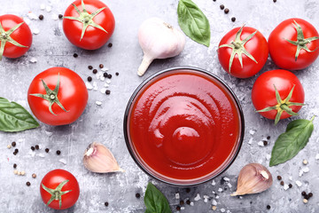 Ketchup in bowl with basil leafs and garlic on grey wooden table