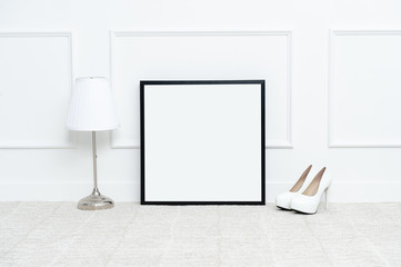 There are beautiful flowers and various objects near the rectangle blank picture frame in front of white wall on the carpet living room.