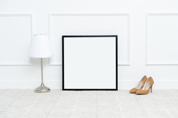 There are beautiful flowers and various objects near the rectangle blank picture frame in front of white wall on the carpet living room.