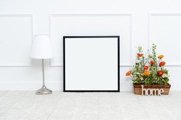 There are beautiful flowers and various objects near the rectangle blank picture frame in front of white wall on the carpet living room.