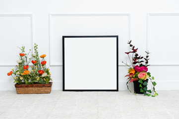 There are beautiful flowers and various objects near the rectangle blank picture frame in front of white wall on the carpet living room.