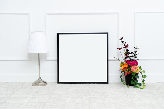 There are beautiful flowers and various objects near the rectangle blank picture frame in front of white wall on the carpet living room.