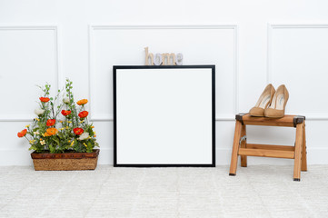 There are beautiful flowers and various objects near the rectangle blank picture frame in front of white wall on the carpet living room.