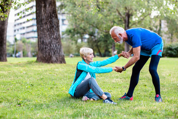 Senior woman helping her husband to stand up after fall while they were jogging.