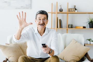 cheerful senior man sitting on sofa and holding remote control while waving hand