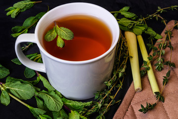Herbal tea in a white mug on a dark background. Fresh herbs around: mint, thyme and lemon sargo.