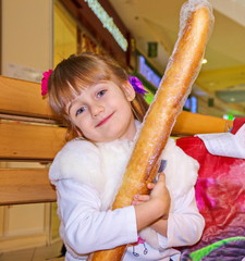 Charming little girl sits on a bench in a store next to a shopping bag and holds a baguette in her hands.