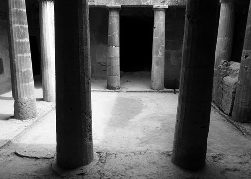 monochrome image of an underground chamber at the tombs of the kings in paphos cyprus with old eroded sandstone columns surrounding a dark empty doorway