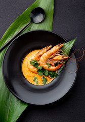 Seafood cream soup with shrimps decorated with tropical leaves. In black plate, on black background. Top view, copy space.