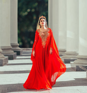 Gorgeous turkish empress shakherezad luxurious red flying dress with long train, sleeves, gold. walks smoothly between large columns with crown, earrings and bright scarlet lipstick. art sexy photo
