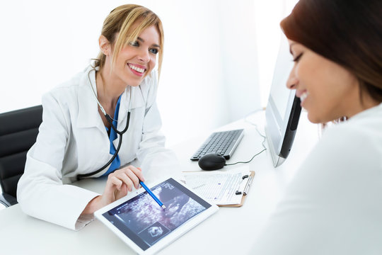 Young woman gynecologist doctor showing to pregnant woman ultrasound scan baby with digital tablet in medical consultation.