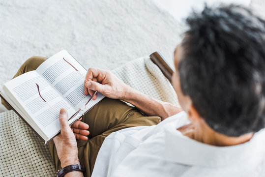 selective focus of senior man reading book and holding glasses at home