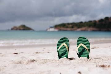 Green flip flops in the white sandy beach near sea waves, nobody. Summer vacation concept with blue...