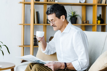 retired man in glasses holding cup with coffee and reading newspaper at home