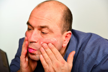 Naklejka premium An adult and unshaven man in a blue shirt applies a mask of cream on his face, the concept of fashion, style and personal care.