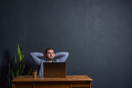 Satisfied with work done. Happy young man working on laptop while sitting at his working place in office Dark background. Space for Text