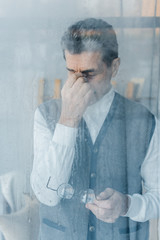 tired senior man touching eyes while standing near window