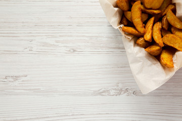 Fried potato wedges in paper box on a white wooden surface. Overhead, top view, from above. Copy space.