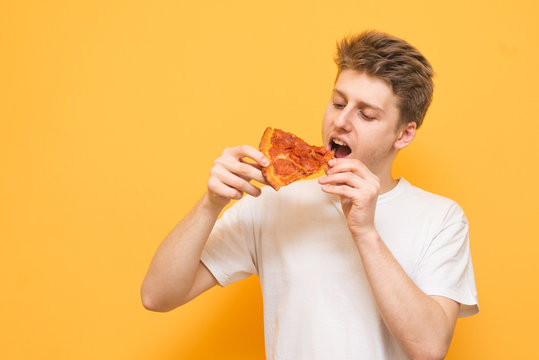 Portrait Of A Young Man Eating A Piece Of Fresh Tasty Pizza On A Yellow Background. Hungry Guy With A Piece Of Pizza In His Hands, Looking At Fast Food And Going To Eat.