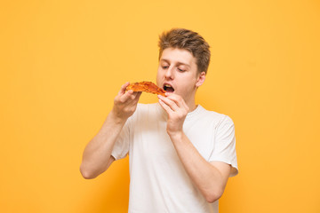 Portrait of a guy in a white T-shirt holds a piece of pizza in his hands, looking and eating. Hungry guy eats fast food, pizza on a yellow background. Copyspace