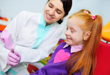 children's dentist examines the teeth and mouth of the child - a cute red-haired girl sitting in a dental chair