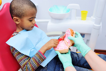 a pediatric dentist teaches an African American child who sits in a dental chair to brush his teeth properly. Pediatric dentistry